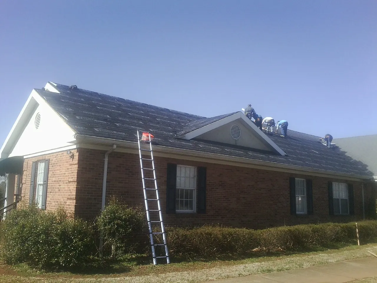 Crew tearing shingles off a brick ranch roof with a ladder set — Metal Roof Repair work by our crew in Malone